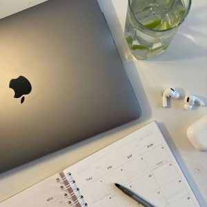 Ordenador mac, un vaso de agua con limón, airpods y agenda con boli