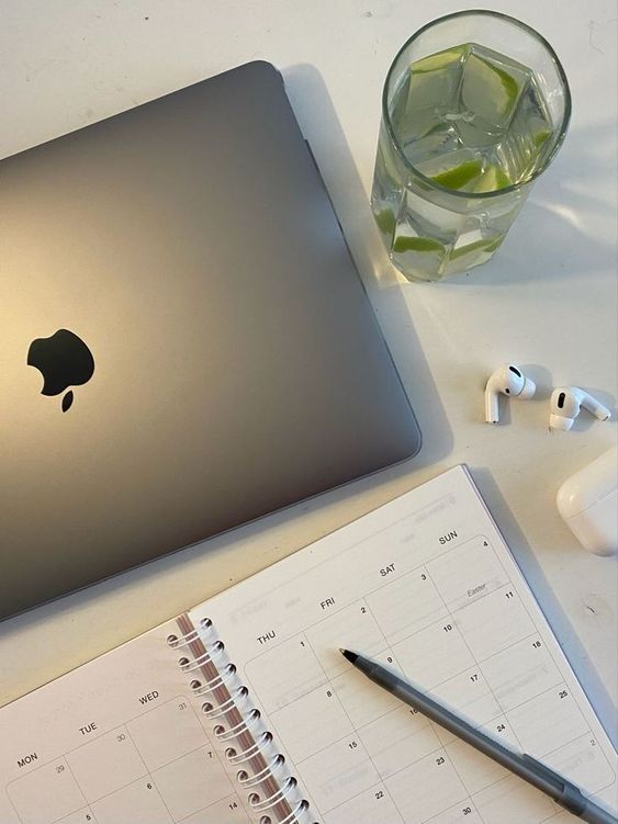 Ordenador mac, un vaso de agua con limón, airpods y agenda con boli